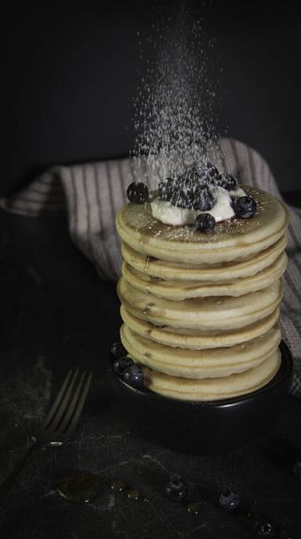 De bovenkant van een stapel pannenkoeken bestrooien met poedersuiker. Yoghurt en bosbessen topping.  - Foto, afbeelding
