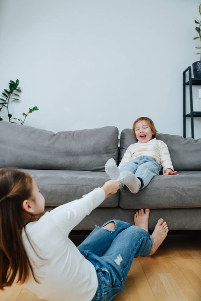 Strong girl making a physical effort, pulling off sock of a boy, they are preparing for a tickling competition. He is sitting on a couch in a living room, he is on the floor. - Foto, imagen