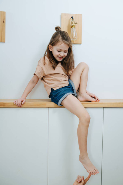 Curious little girl sitting on a corridor shelf giving her foot to be tickled, showing off, being brave. Having fun. - Foto, Bild