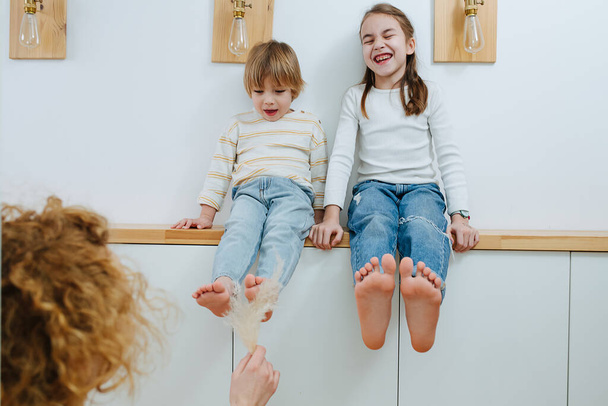 Mom tickling her children's feet with a fluffy cereal spikes. They are sitting on a shelf, a girl is laughing, a boy is slightly afraid. - Photo, image