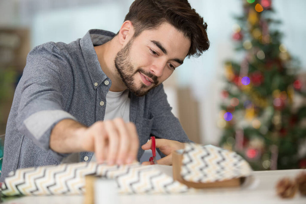 young man wrapping gifts and making bow on table - Photo, Image
