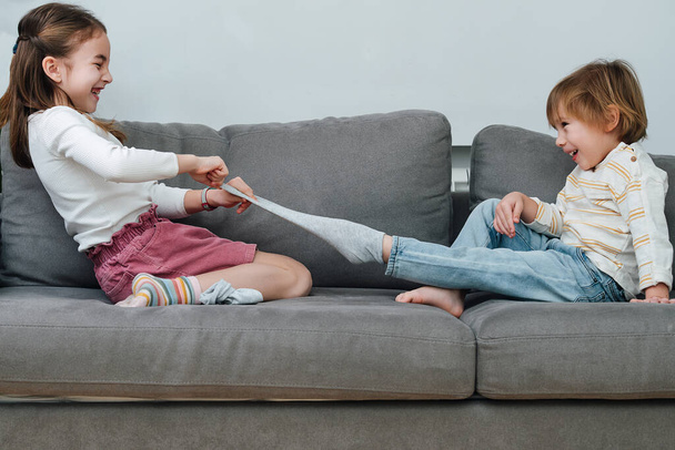 Laughing girl pulling off sock of a boy, they are preparing for a tickling competition. Sitting on a couch in a living room. Side view. - 写真・画像