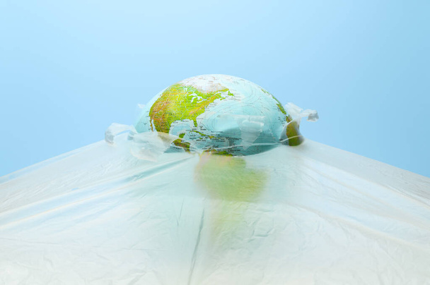 El globo de la tierra, cubierto con una envoltura de plástico sobre un fondo azul pastel. Día de la Tierra, Día Mundial del Medio Ambiente. Concepto de contaminación planetaria con polietileno de celofán plástico - Foto, imagen