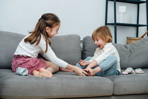 Happy girl trying to tickle boy's foot, they both heartedly laugh. On a grey couch in the living room. - Фото, изображение