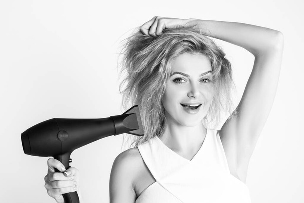 Health hair and beauty concept. Girl with blonde hair using hairdryer. Young attractive happy laughing blonde woman with hair dryer. Hairstyle, hairdressing concept. - Фото, изображение