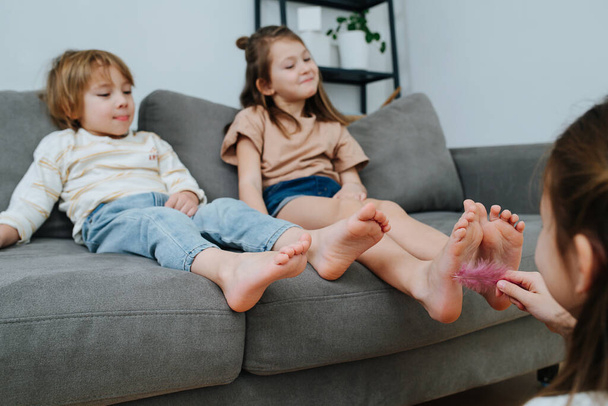 Girl conducting tickling competition between siblings sitting on a couch using a feather. Their faces blurred, focus on feet. - Фото, изображение