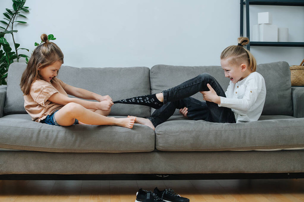 Girl pulling off sock of a boy, they are preparing for a tickling competition. Sitting on a couch in a living room. Side view. - 写真・画像