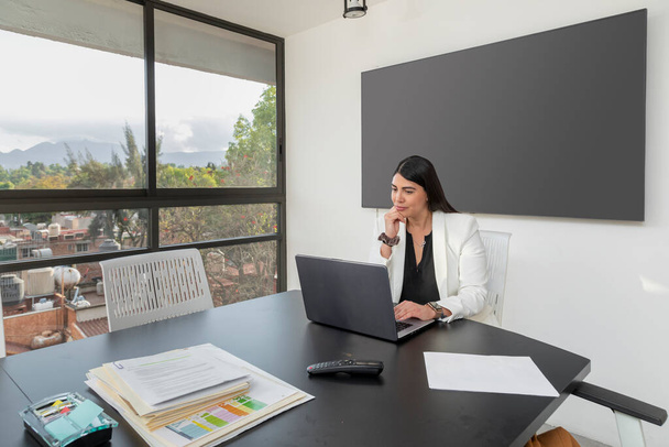 Verveelde zakenvrouw werkzaam in de directiekamer van haar kantoor, met een scherm voor presentatie aan de muur en ramen met uitzicht naar buiten. - Foto, afbeelding