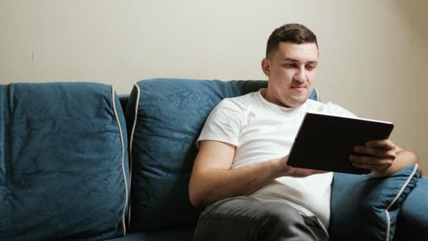 30-letni mężczyzna o europejskim wyglądzie siedzi na ciemnoniebieskiej kanapie i pracuje na tablecie. Facet uśmiecha się patrząc na ekran tabletu. - Materiał filmowy, wideo