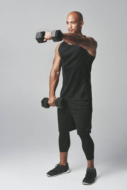 Working on his upper body. Studio shot of an athletic young man working out with dumbbells against a grey background. - Foto, afbeelding
