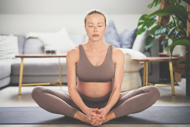 Young beautiful pregnant woman practising yoga at home in her living room. Motherhood, pregnancy, healthy lifestyle, well being and yoga concept. - Φωτογραφία, εικόνα