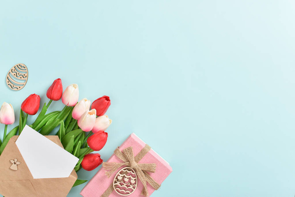 A bouquet of tulips with a postcard and a gift and wooden Easter eggs on a light blue background. Happy Easter. - Photo, Image