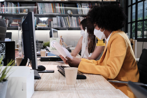 新しい通常オフィスで働くフェイスマスクを持つ2人の女性同僚チーム。COVID-19クリアパーティション、事業場オフィス、パンデミック健康のための社会的距離、病気の予防による保護. - 写真・画像