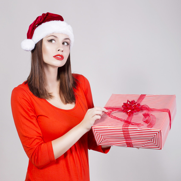Brunette Woman in Red Dress and Santa Hat holding Present and Looking in camera - Photo, Image