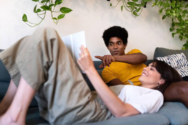 Nuori rotujenvälinen pari rentoutua yhdessä kotona. Makaa sohvalla lukemassa kirjaa. - Valokuva, kuva