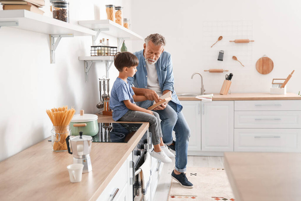 Senior man reading book to his little grandson in kitchen - Foto, Imagem
