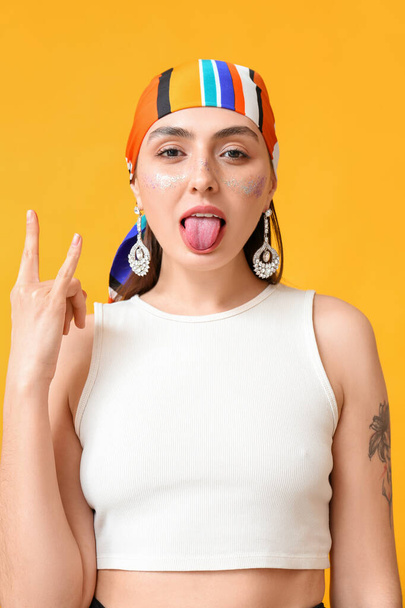 Cool femme à la mode avec maquillage créatif montrant la langue sur fond jaune - Photo, image
