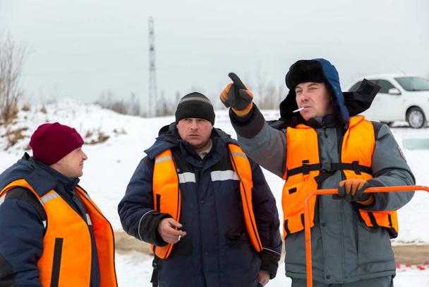 Pracovníci montéři v oranžových záchranných vestách na ledě zamrzlého jezera na kouřové přestávce - Fotografie, Obrázek