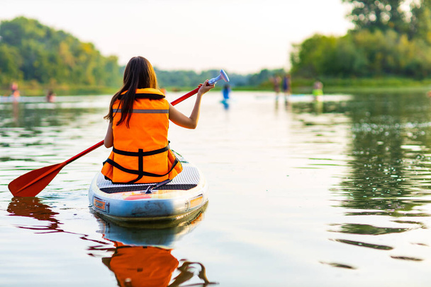young woman in orange life jacket on supboard at river - Foto, imagen