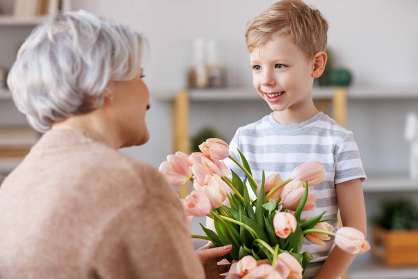 Cheerful little boy giving flowers to grandmother - Фото, изображение