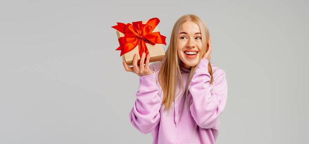 Portrait de Happy jeune femme blonde en sweat à capuche tendance tenant boîte cadeau avec ruban rouge et en regardant l'appareil photo sur fond gris. Nouvel An Fête des femmes anniversaire concept de vacances. Copiez l'espace pour votre texte - Photo, image