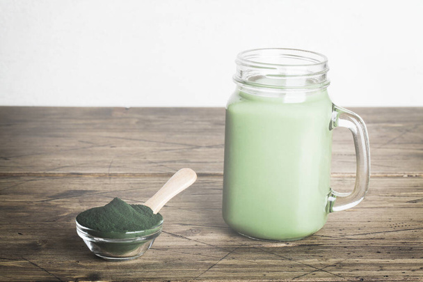 Cam şişede taze yeşil smoothie ve ahşap arka planda kaşıkla spirulina tozu. kullanışlı alışkanlıklar - Fotoğraf, Görsel