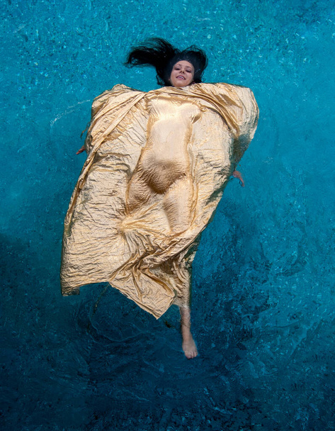 nainen kelluu makaa selällään turkoosi vesi altaan peitetty kultainen kangas, jonka alla hänen seksikäs kehon muodot ovat kauniisti hahmoteltu, kopioi tilaa, - Valokuva, kuva