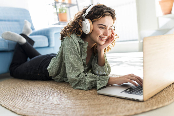 lockige Frau, die zu Hause mit Laptop auf dem Teppich liegt, Musik hört, studiert. - Foto, Bild