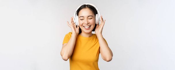 Boldog ázsiai lány tánc, zenét hallgatni a fejhallgató és mosolygós, álló sárga póló ellen fehér háttér - Fotó, kép
