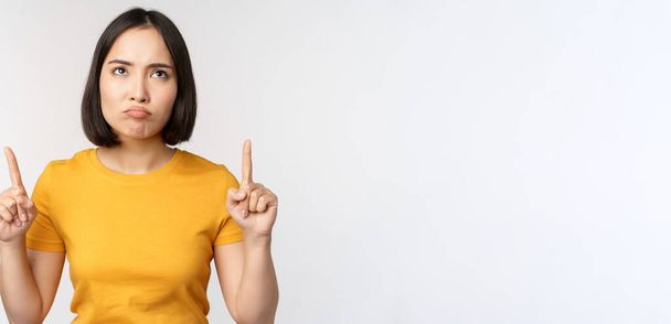 Hüsrana uğramış Asyalı kadın bakıyor, kızgın suratıyla parmaklarını kaldırıyor, beyaz arka planda sarı tişörtle dikiliyor. - Fotoğraf, Görsel