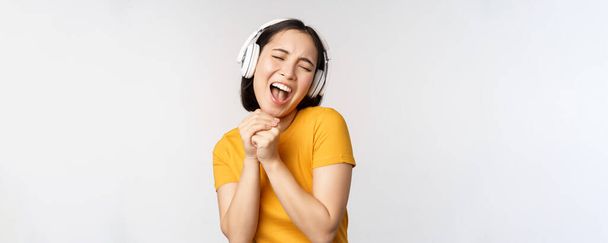 Heureuse fille asiatique dansant, écoutant de la musique sur écouteurs et souriant, debout en tshirt jaune sur fond blanc - Photo, image
