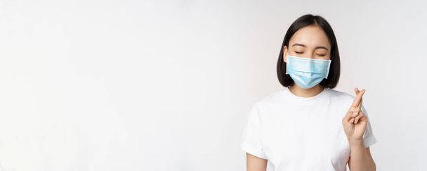 Covid-19, healthcare and medical concept. Image of asian girl in medical face mask, cross fingers, praying, making wish and smiling, standing over white background - Foto, immagini