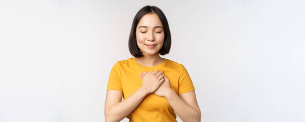 Romantický asijské přítelkyně, držící se za ruce na srdci, usmívající se s péčí a něhou, stojící ve žluté tričko přes bílé pozadí - Fotografie, Obrázek