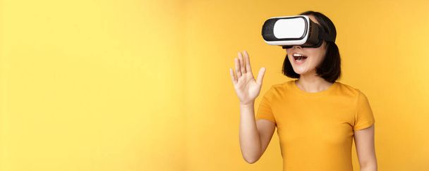 Une fille en VR. Belle jeune femme asiatique, en utilisant des lunettes de réalité virtuelle et en jouant, bavardant virtuellement, debout sur fond jaune - Photo, image