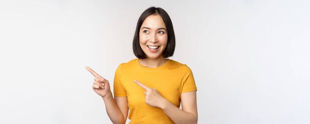 Portrait de fille brune asiatique souriante en tshirt jaune, pointant les doigts vers la gauche, montrant l'espace de copie, promo deal, démonstration de bannière, debout sur fond blanc - Photo, image