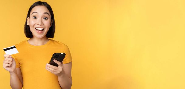 Verkkokaupoissa. Iloinen aasialainen tyttö, jolla luottokortti ja älypuhelin, maksaa, tilaus matkapuhelimella, seisoo keltaisen taustan päällä - Valokuva, kuva