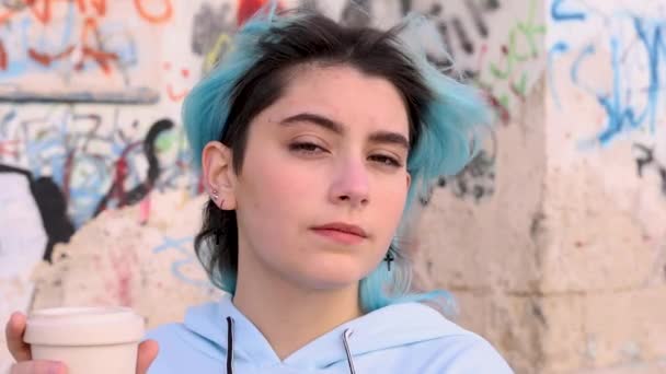 Tinédzser lány kék, túlméretezett kapucnis pulcsiban kávét iszik elvitelre. Kék hajú tinilány kint marad a graffiti fal mellett. Mockup, fejlövés. Hipszter és serdülőkor koncepció - Felvétel, videó