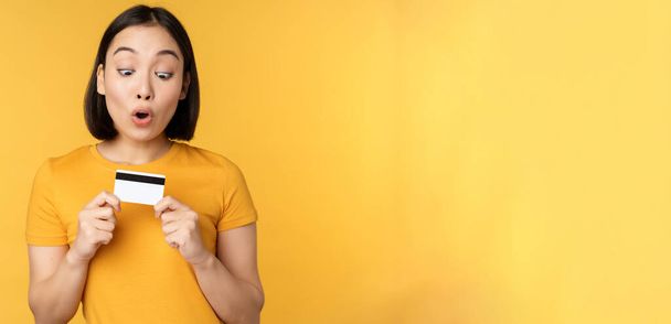 Beautiful asian woman showing credit card and smiling, recommending bank service, standing over yellow background - Fotó, kép