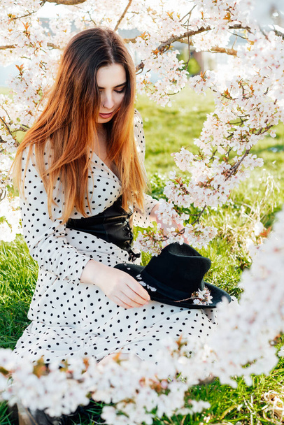Young brunette woman in dress with hat sitting near a white blooming tree and enjoying the moment. Generation Z girl. Spring mood. Relax on the nature. Vertical card. - Foto, immagini
