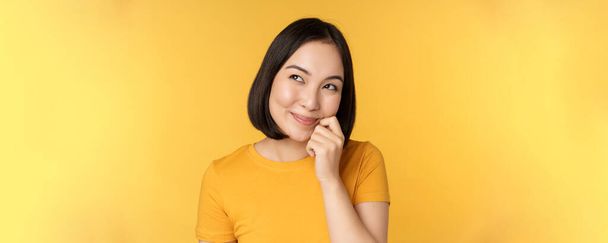 Primer plano retrato de linda chica asiática sonriendo, pensando, mirando hacia arriba reflexivo, de pie en camiseta sobre fondo amarillo - Foto, Imagen