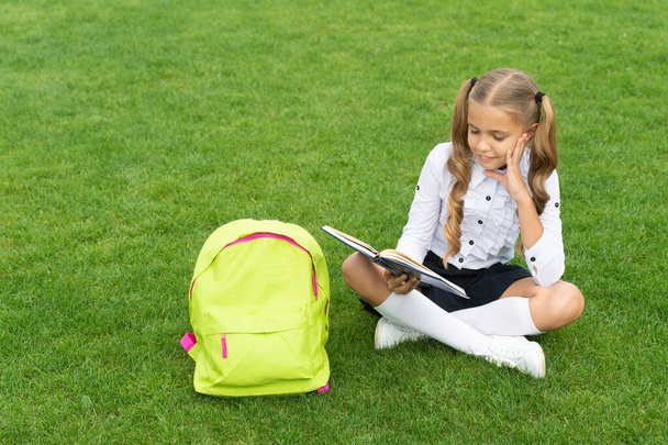 Happy child in uniform with school bag reading book sitting on green grass - Фото, зображення