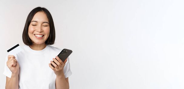 Verkkokaupoissa. Onnellinen aasialainen nainen, joka käyttää luottokorttia ja älypuhelinsovellusta, maksaa verkkosivuilla matkapuhelimen kautta, valkoinen tausta - Valokuva, kuva