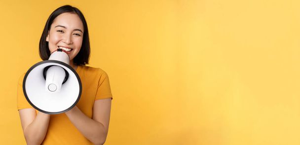 Mosolygó ázsiai nő áll megafon, bejelentette smth, reklám termék, áll a sárga háttér - Fotó, kép