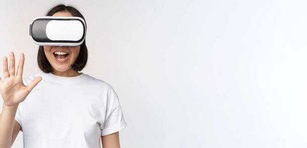 Gelukkig aziatische vrouw met behulp van VR-headset, zwaaien opgeheven handen en lachen, met behulp van virtual reality bril, staande over witte achtergrond - Foto, afbeelding