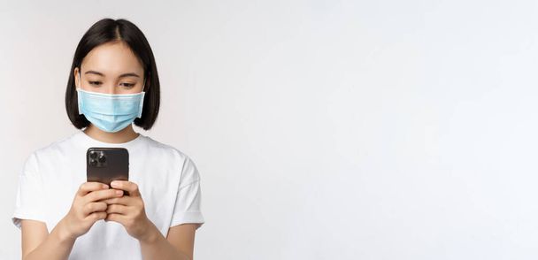 Concepto de salud, covidio y móvil. Mujer asiática joven en máscara facial médica, mirando la pantalla del teléfono inteligente, utilizando la aplicación del teléfono, compras en línea, fondo blanco - Foto, Imagen