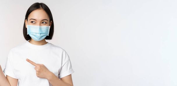 Mujer coreana joven con máscara facial médica apuntando con los dedos hacia la izquierda y mirando el logotipo, mostrando publicidad o pancarta, de pie sobre fondo blanco - Foto, Imagen