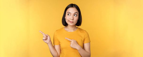 Portret zaintrygowanej Azjatki, patrzącej i wytykającej palcami w lewo na reklamę, pokazującej smth ciekawy, stojący nad żółtym tłem - Zdjęcie, obraz