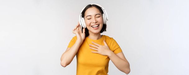 Χαρούμενη Ασιάτισσα που χορεύει, ακούει μουσική με ακουστικά και χαμογελάει, στέκεται με κίτρινο μπλουζάκι σε λευκό φόντο - Φωτογραφία, εικόνα