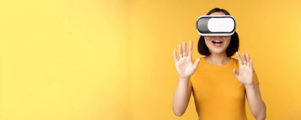 Young asian woman playing in VR, using virtual reality glasses, wearing yellow t-shirt against studio background - 写真・画像