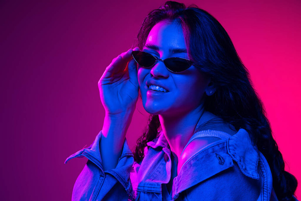 Beautiful young emotional girl, student with long dark hair isolated on gradient pink-blue background in neon light. Concept of emotions, youth - 写真・画像